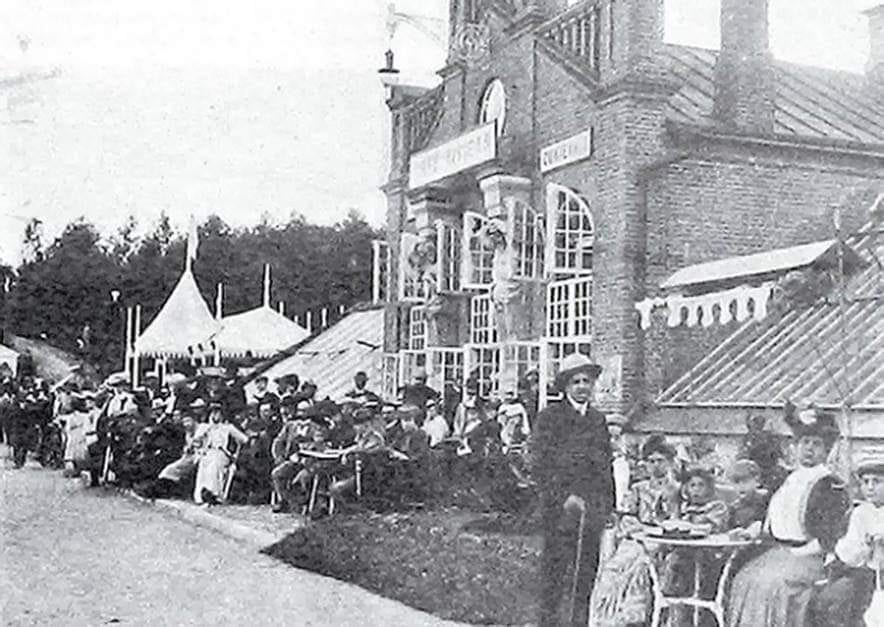 Старая фотография: публика за столиками у кирпичного здания кафе с террасой, рядом белый шатёр и лес